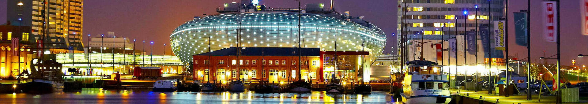 Bremerhaven bei Nacht mit beleuchtetem ATLANTIC Hotel Sail City am Wasser, umgeben von Gebäuden und Booten.