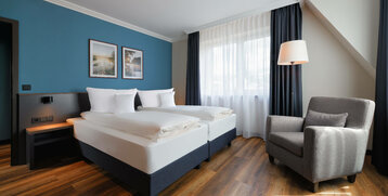 Modernes Hotelzimmer mit Doppelbett, blauer Wand, zwei Bildern, Sessel und großem Fenster mit Vorhängen.