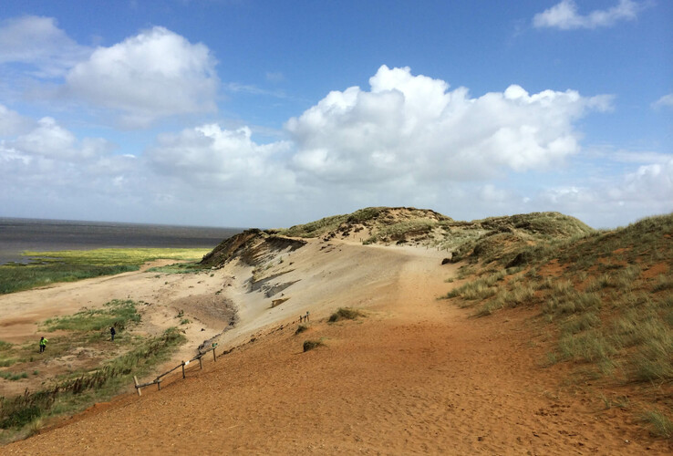 Sand dune landscape with blue sky and scattered clouds, the sea in the background.