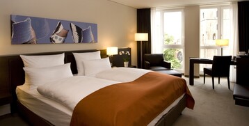 Modernes Hotelzimmer mit Doppelbett, orangefarbener Decke, Schreibtisch, Sesseln und großem Fenster mit Stadtblick.