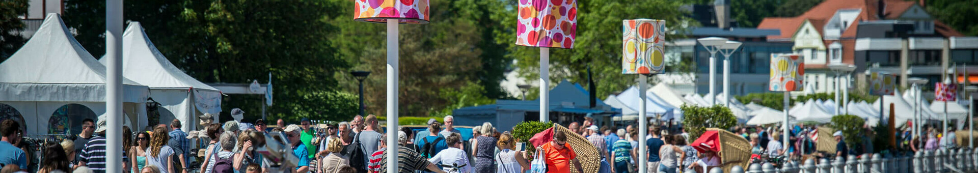 Promenade in Travemünde mit Menschen, bunten Laternen und Zelten, bei sonnigem Wetter.