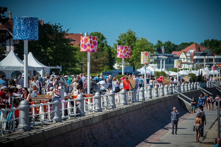 Promenade in Travemünde mit Menschen, bunten Laternen und Zelten, bei sonnigem Wetter.