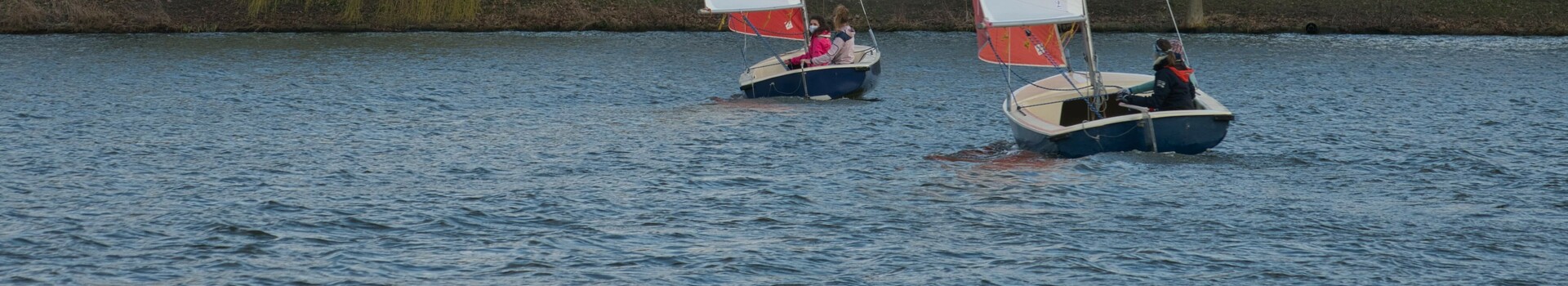 Angebot Segeln in Münster l ATLANTIC Hotel Münster Zwei Segelboote auf einem See vor einer Baumreihe und Häusern, mit ATLANTIC Hotel Münster Logo auf den Segeln.
