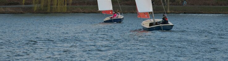 Zwei Segelboote auf einem See vor einer Baumreihe und Häusern, mit ATLANTIC Hotel Münster Logo auf den Segeln.