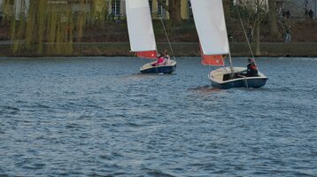 Zwei Segelboote auf einem See vor einer Baumreihe und Häusern, mit ATLANTIC Hotel Münster Logo auf den Segeln.