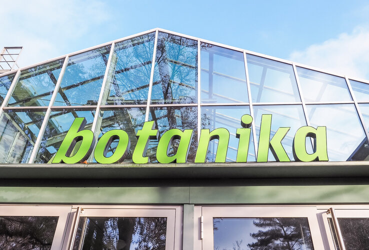 Glasfassade mit grünem Schriftzug "botanika", Bäume spiegeln sich im Glas.