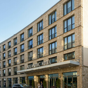 Fassade des ATLANTIC Hotels Lübeck mit moderner Architektur, Eingang und davor geparktem Auto bei sonnigem Wetter.