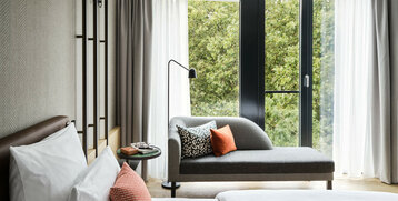 Helles Hotelzimmer mit Bett, grauem Sofa, bunten Kissen und großen Fenstern mit Blick auf grüne Bäume.