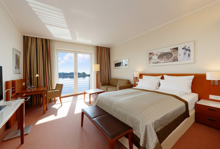 Helles Hotelzimmer mit Doppelbett, Sofa, Schreibtisch und großem Fenster mit Seeblick.