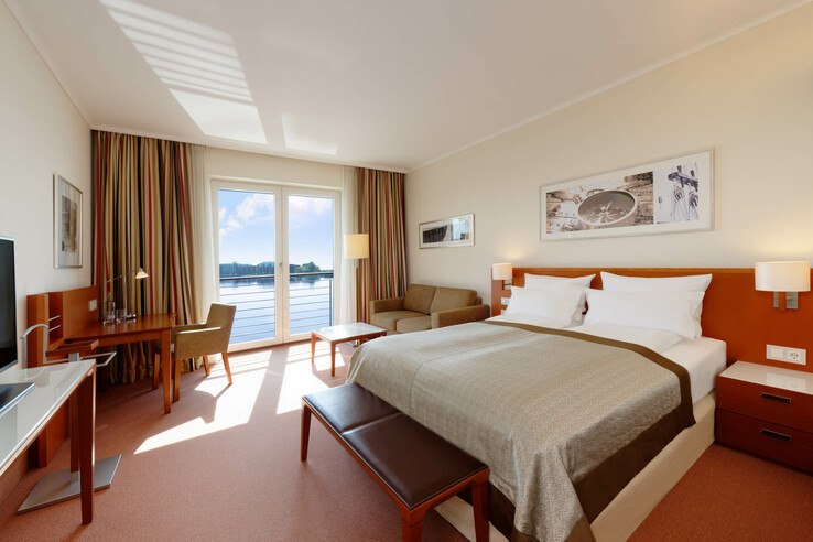 Helles Hotelzimmer mit Doppelbett, Sofa, Schreibtisch und großem Fenster mit Seeblick.