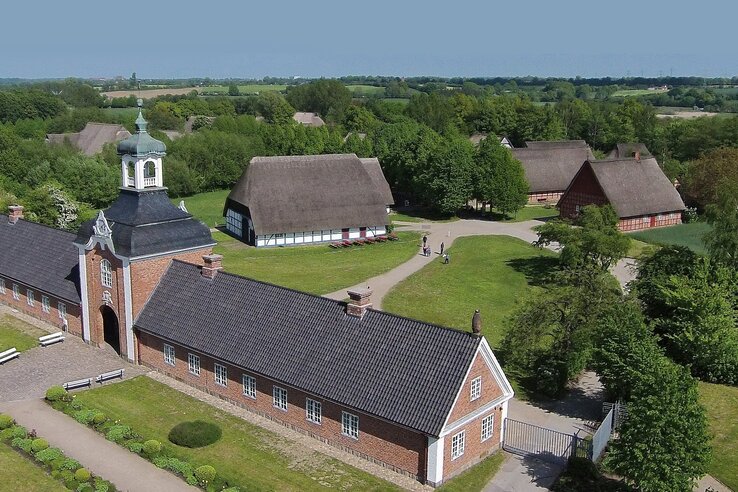 Historic brick building with tower, surrounded by green countryside and traditional thatched roof houses.