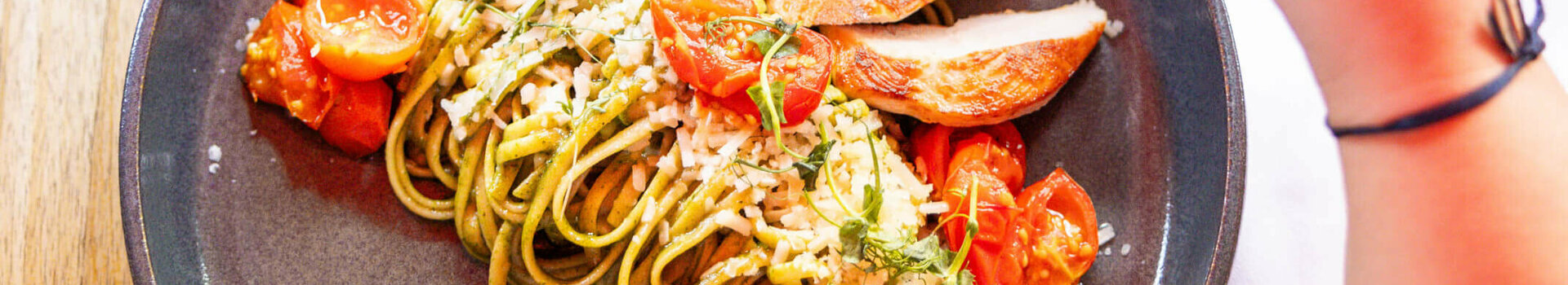 Plate with pasta, grilled chicken, cherry tomatoes and herbs, hand holding a glass of wine.