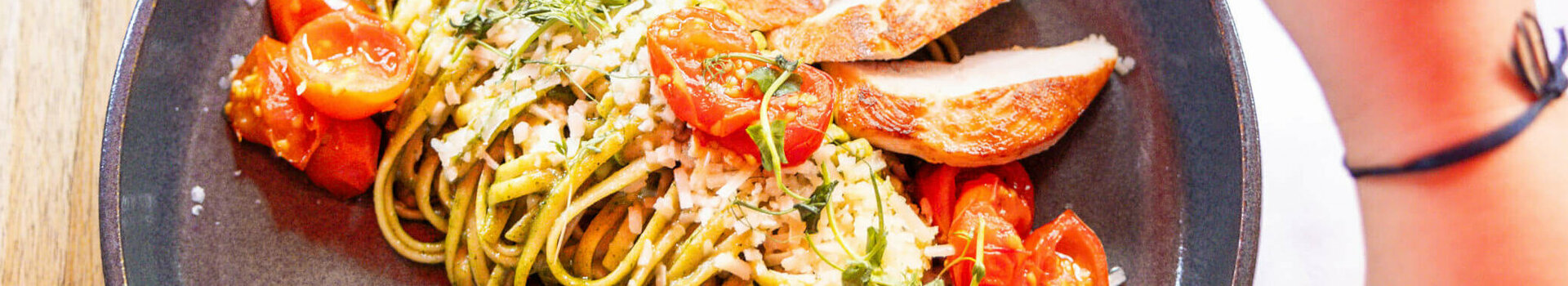 Teller mit Pasta, gegrilltem Hähnchen, Kirschtomaten und Kräutern, Hand hält ein Glas Wein.