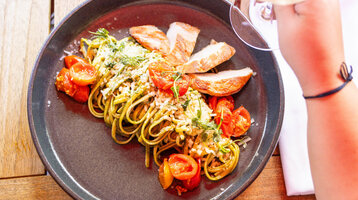 Teller mit Pasta, gegrilltem Hähnchen, Kirschtomaten und Kräutern, Hand hält ein Glas Wein.