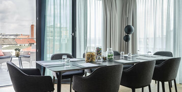 Modern conference room with large table, chairs and glasses, bright curtains, balcony with city view in the background.