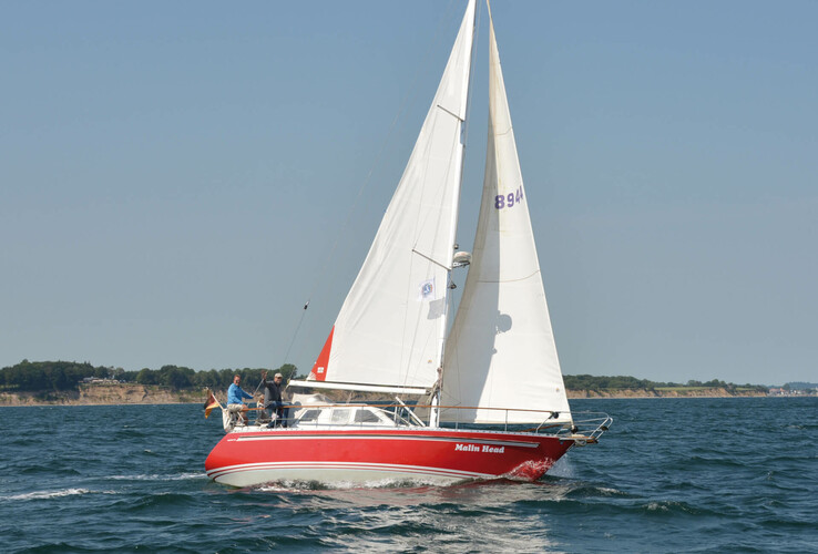 Segelboot mit weißen Segeln und rotem Rumpf auf der Ostsee, Küste im Hintergrund, blauer Himmel.