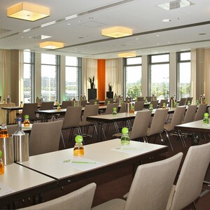Modern conference room with rows of tables and chairs, equipped with drinks and writing materials, large windows.