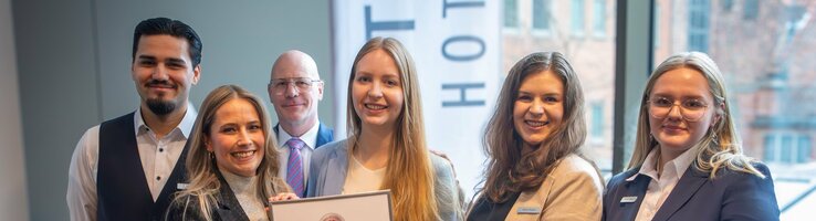 Eine Gruppe von sechs Hotelmitarbeitern hält stolz ein Zertifikat in einem modernen Raum.