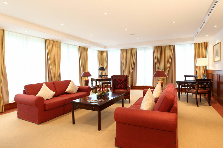 Helles Wohnzimmer mit roten Sofas, Holztisch, Schreibtisch und großen Fenstern mit goldenen Vorhängen.