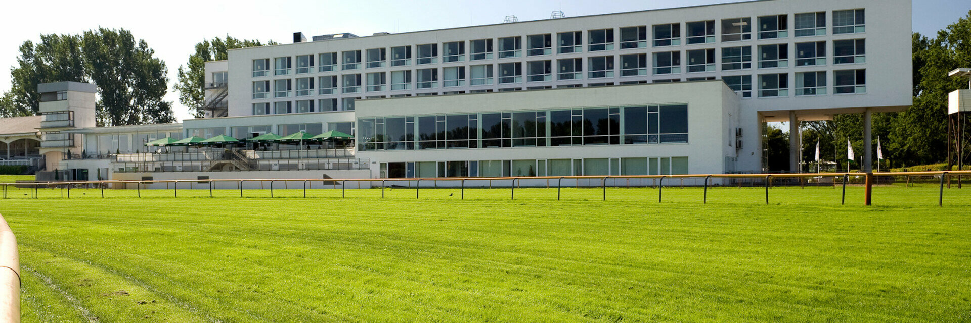 ATLANTIC Hotel Galopprennbahn, modernes Gebäude mit großen Fenstern, umgeben von grünem Rasen und Bäumen.