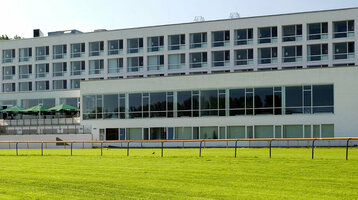 ATLANTIC Hotel Galopprennbahn, modernes Gebäude mit großen Fenstern, umgeben von grünem Rasen und Bäumen.