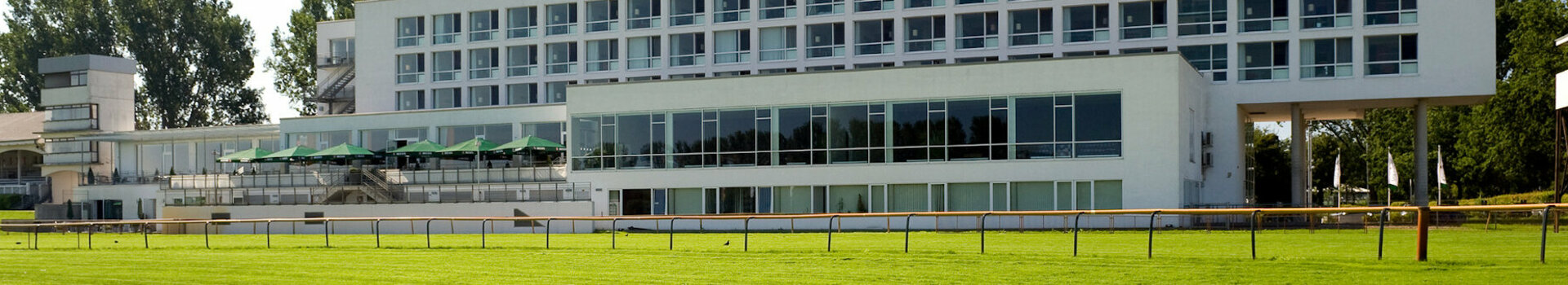 ATLANTIC Hotel an der Galopprennbahn ATLANTIC Hotel Galopprennbahn, modernes Gebäude mit großen Fenstern, umgeben von grünem Rasen und Bäumen.