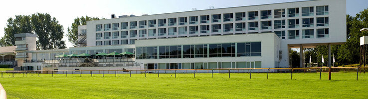ATLANTIC Hotel Galopprennbahn, modernes Gebäude mit großen Fenstern, umgeben von grünem Rasen und Bäumen.