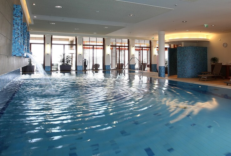 Helles Hallenbad mit Wasserfall, Liegestühlen und großen Fenstern, die viel Tageslicht hereinlassen.