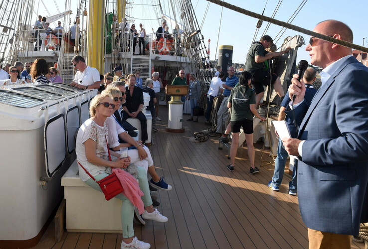 Person hält Rede auf einem Segelschiff, umgeben von sitzenden und stehenden Zuhörern.