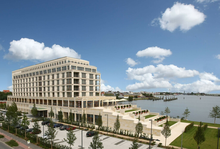 Modernes Hotel am Wasser mit großen Fenstern, Terrasse, gepflegter Gartenanlage und Parkplatz, unter blauem Himmel.