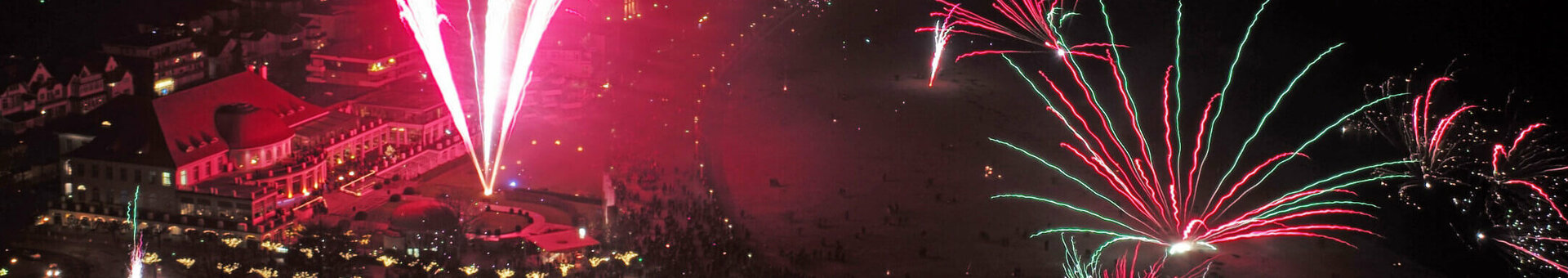 Neujahrsgarten mit großem Silvesterfeuerwerk Feuerwerk über dem ATLANTIC Grand Hotel Travemünde bei Nacht, mit Menschenmengen und beleuchteten Zelten im Vordergrund.