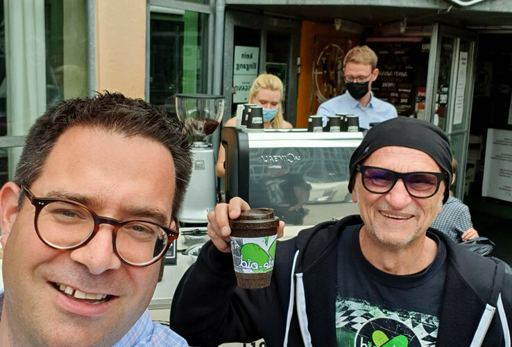 Two men smiling in front of a mobile coffee stand, one holding an organic coffee cup. Barista with mask in the background.