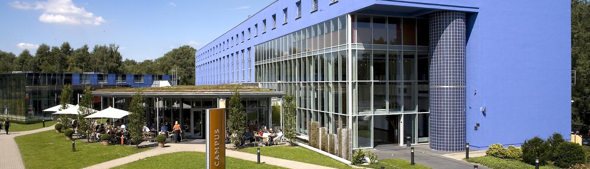 Blaues Hotelgebäude mit Glasfassade, grüner Rasen, Terrasse mit Gästen und Sonnenschirmen, sonniger Tag.