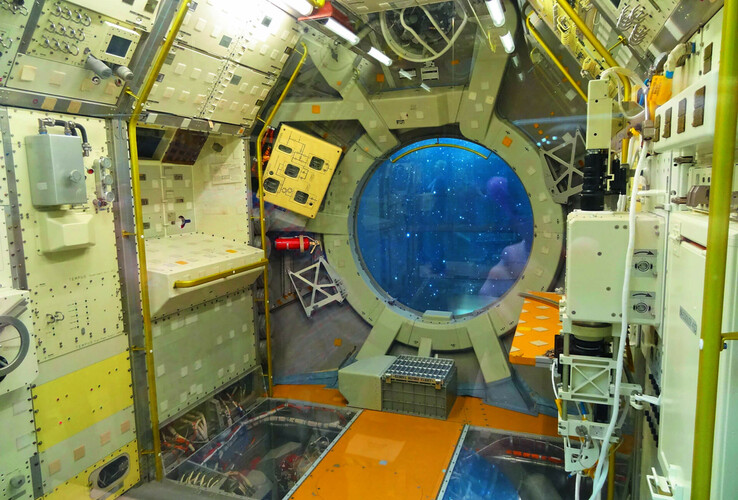 Interior of a space station with technical equipment and a large round window showing the view into space.