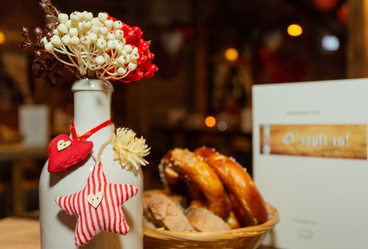 Dekorative Vase mit roten und weißen Blumen, Herz- und Sternanhänger. Daneben Brezeln in einem Korb, festliche Atmosphäre.