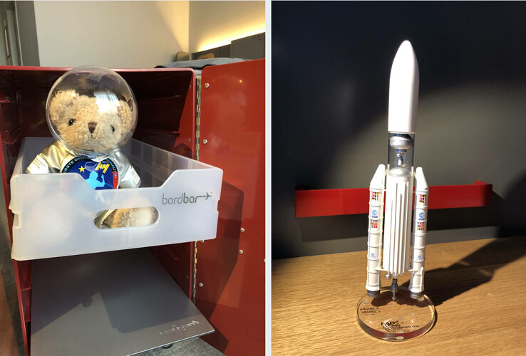 Teddybär in Astronautenanzug in Bordbar, daneben Modell einer Ariane 5 Rakete auf Holzregal.
