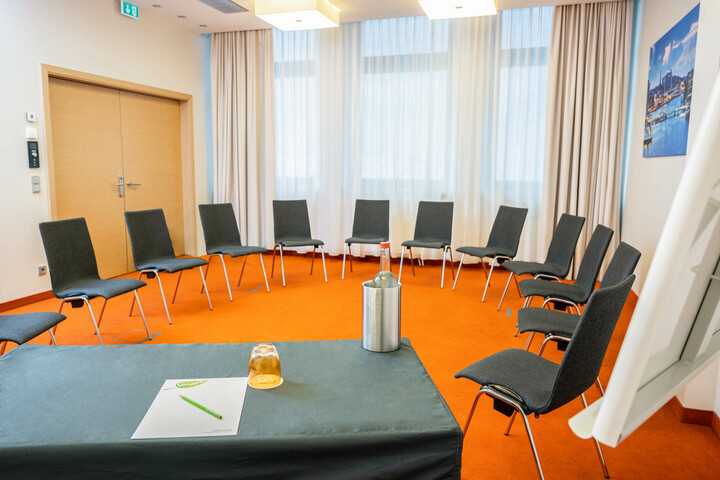 Konferenzraum mit Stuhlkreis, Tisch mit Wasserflasche, Glas und Schreibmaterial, orangefarbener Teppich.