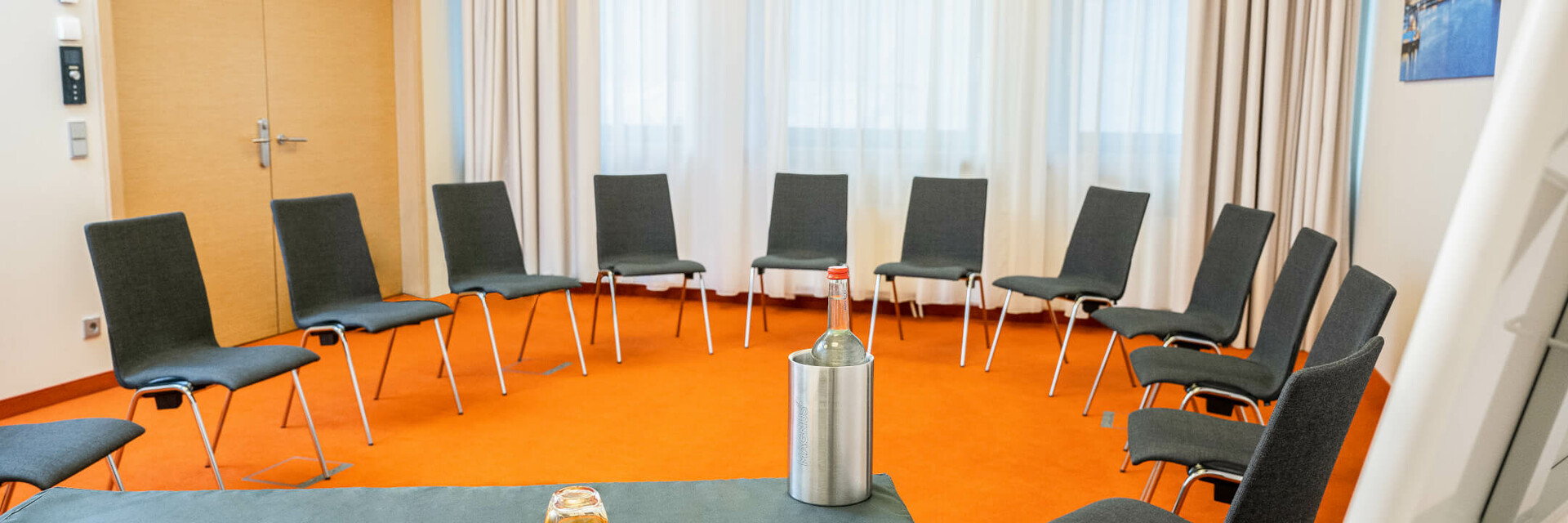 Konferenzraum mit Stuhlkreis, Tisch mit Wasserflasche, Glas und Schreibmaterial, orangefarbener Teppich.