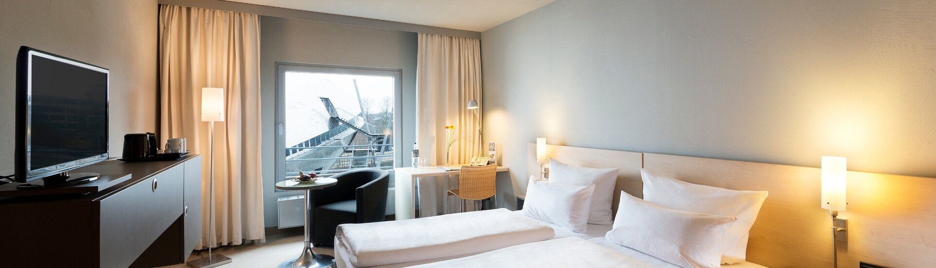 Innenansicht des Comfort Zimmers mit gemütlicher Ausstattung im ATLANTIC Hotel Universum Bremen Helles Hotelzimmer mit Doppelbett, Schreibtisch, TV und Blick auf moderne Architektur durch großes Fenster.