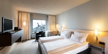 Bright hotel room with double bed, desk, TV and view of modern architecture through large window.