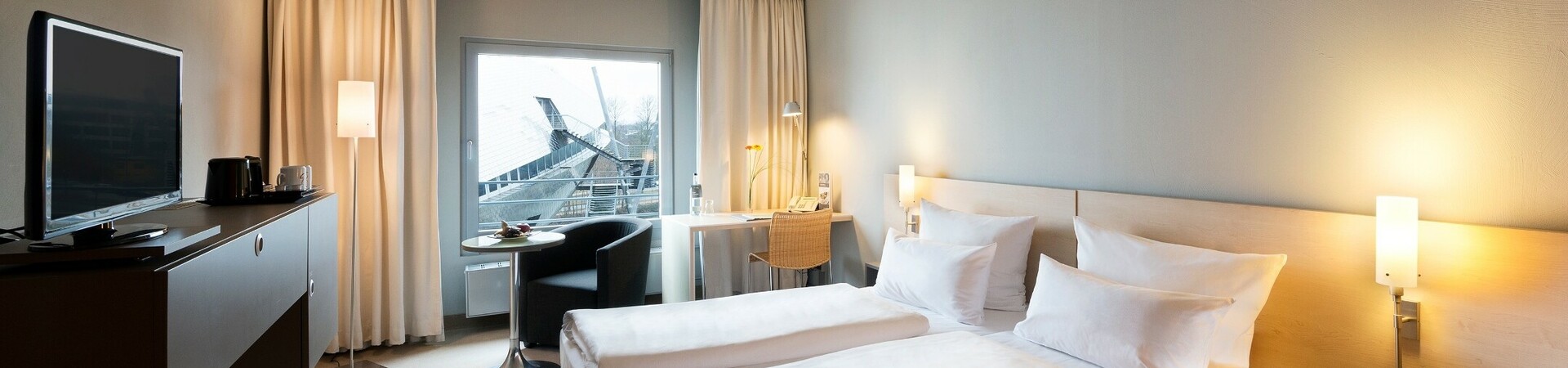 Helles Hotelzimmer mit Doppelbett, Schreibtisch, TV und Blick auf moderne Architektur durch großes Fenster.