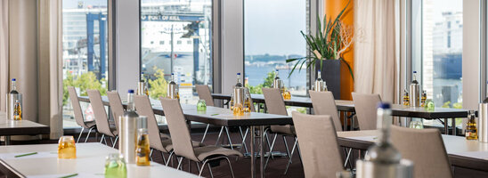 Bright conference room at the ATLANTIC Hotel Kiel with tables, chairs, drinks and a view of the harbor.