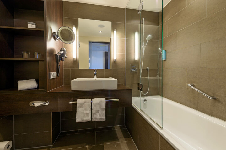 Modernes Badezimmer im ATLANTIC Hotel Lübeck mit Waschbecken, Spiegel, Badewanne und Handtüchern.