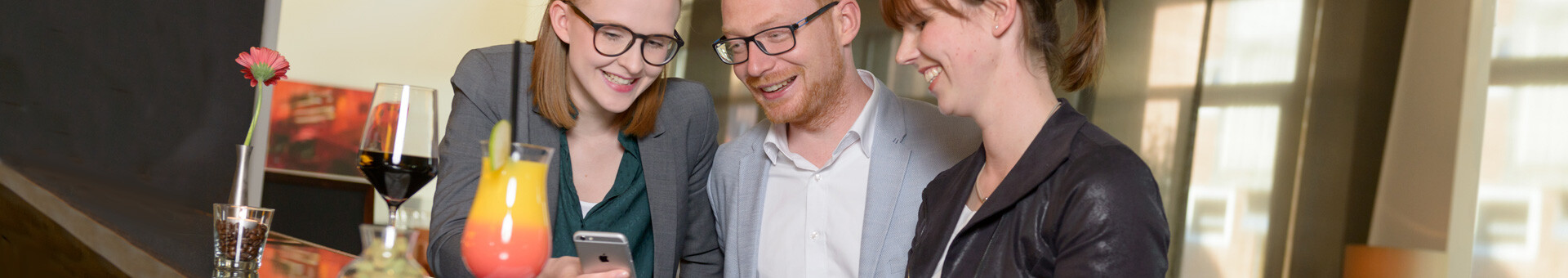 Drei Personen lachen an einer Hotelbar, Getränke und eine Blume stehen auf dem Tresen.