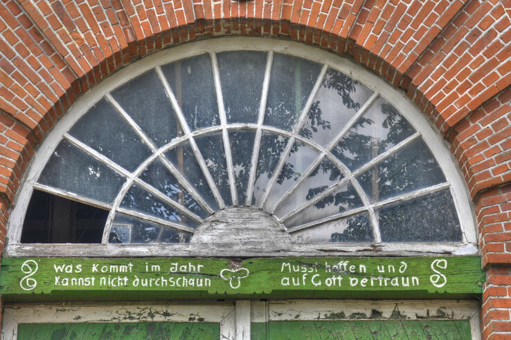 Altes Backsteingebäude mit rundem Fenster und Spruch: „Was kommt im Jahr, kannst nicht durchschau’n. Musst hoffen und auf Gott vertrau’n.“