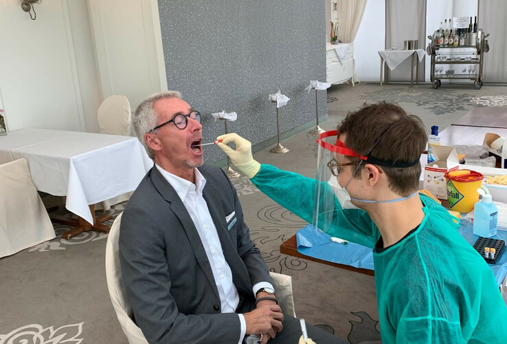 Person in Schutzkleidung führt einen Covid-Test bei einem sitzenden Mann in einem eleganten Hotelraum durch.