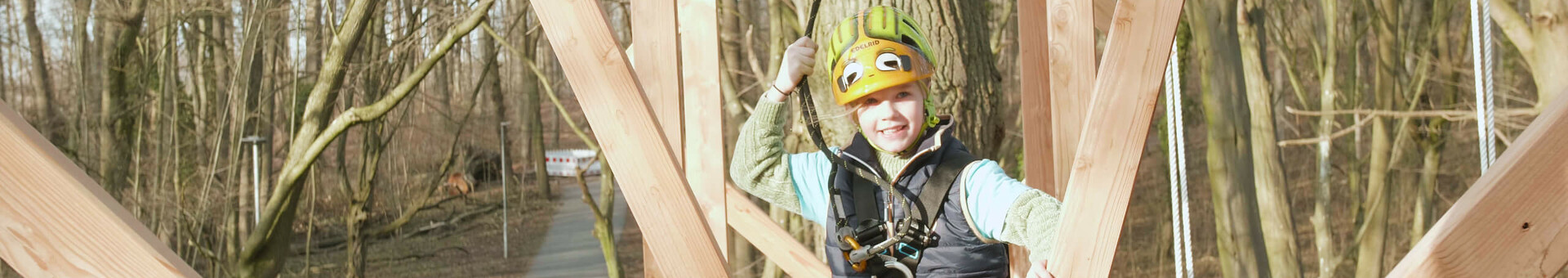 Kletterwald in Travemünde Kind in Kletterausrüstung auf Hochseilgarten-Parcours im Wald, lächelnd und gesichert mit Helm und Gurt.
