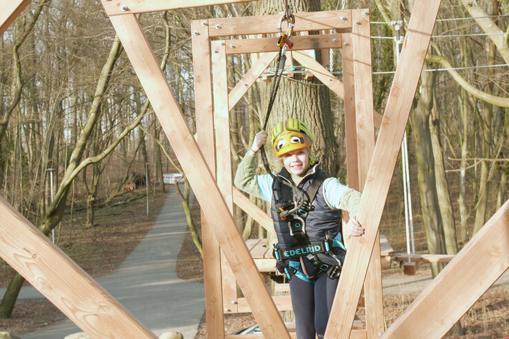 Kind in Kletterausrüstung auf Hochseilgarten-Parcours im Wald, lächelnd und gesichert mit Helm und Gurt.