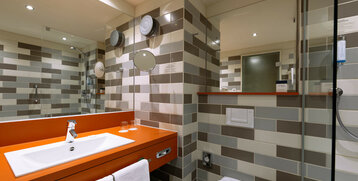 Modern hotel bathroom with orange washbasin, mirror, shower cubicle and towel rail, tiled in shades of gray and beige.