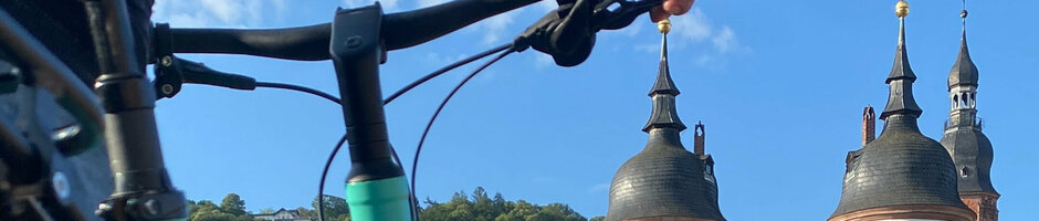 Fahrrad vor historischem Stadttor in Heidelberg mit grüner Landschaft und blauem Himmel im Hintergrund.